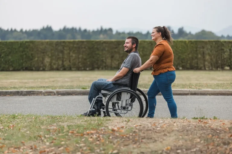 Disabled man in wheelchair being pushed down footpath by support worker, both smiling