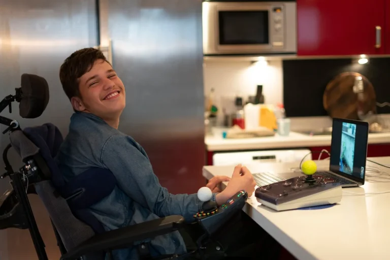 Happy disabled man in wheelchair sitting at kitchen bench looking at laptop