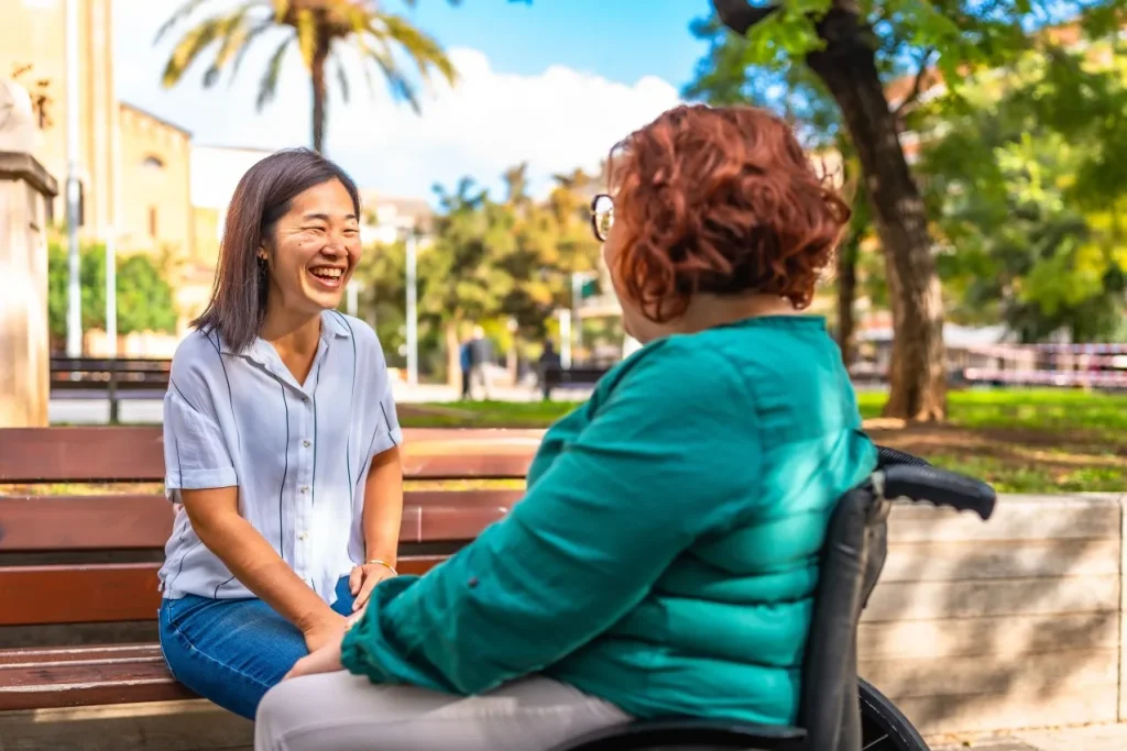 Happy support worker sitting on park bench and laughing with disabled woman in wheelchair